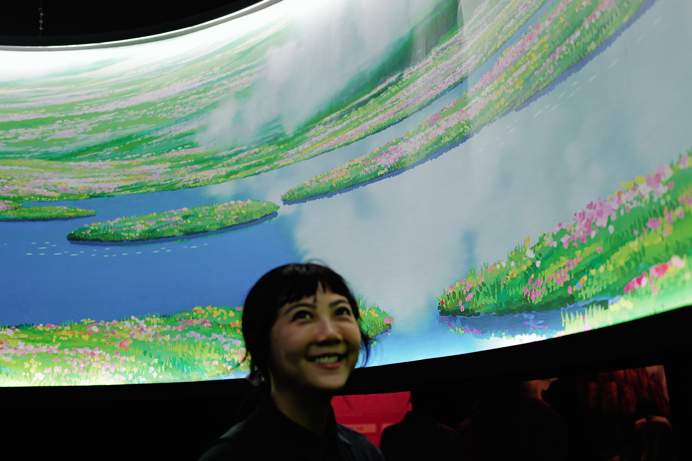 Sijia smiling while looking up at a colorful digital art installation with vibrant greens, blues, and pink abstract patterns